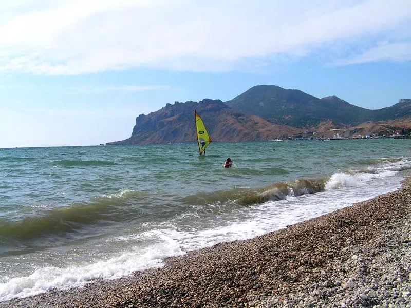 Windsurfer mit gelbem Segel vor einer Küste mit Wellen und Kiesstrand